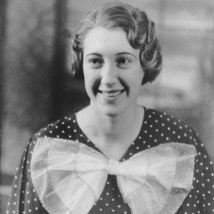 White woman short curled hairstyle wearing polka dot dress with large bow in center smiling
