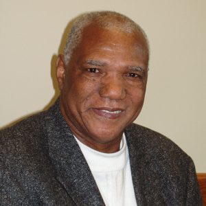 Older black man in sweater and suit jacket wearing name tag text "Arkansas State University Delta Blues Symposium XII Sterling Plumpp"