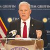 Older white man clean shaven dressed in suit holding glasses speaking at lectern