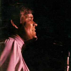 White man short hair pink shirt eyes closed standing in front of microphone