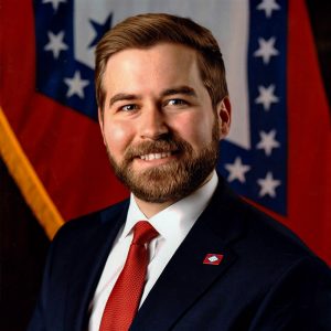 Bearded white man wearing suit and tie with Arkansas state pin attached to suit jacket smiling in front of Arkansas flag