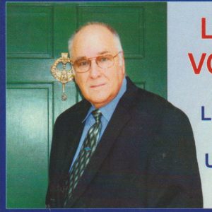 Older white man with receding hairline wearing glasses suit and tie standing in front of door text "Live Green Vote Green Lewis Kennedy for U.S. Congress"