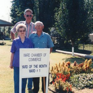 Tall older white man in sunglasses standing behind older white man and woman with wooden sign text "Ward Chamber of Commerce Yard of the Month Ward #1" next to flower bed
