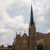 View from parking lot front of multi story brick cathedral tall steeple on right side marble sculpture of man on ground to left of concrete steps leading to double door entrance