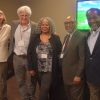 Five individuals standing in row in office building starting on left white woman in suit older white man wearing glasses jeans and suit jacket older black woman in business casual wear middle aged black man wearing glasses and suit and older black man dressed in suit