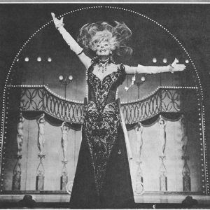 White woman wearing floor length decorative gown necklace headdress and evening gloves standing on stage above text "Carol Channing in Hello