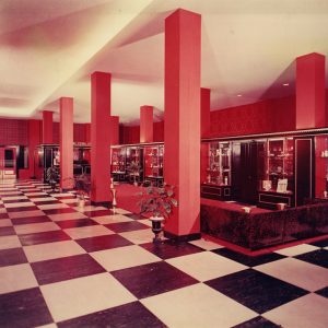 Room with rows of four pillars checkered floors baroque pattern wallpaper couches potted plants and other items in front of several glass display cases along back wall