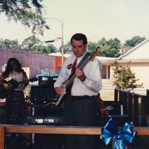 White man wearing dress shirt tie slacks and campaign sticker holding guitar playing on stage next to sound equipment white woman playing guitar wearing sunglasses behind him