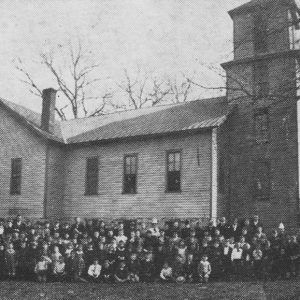 Large group of students front row sitting the rest standing all in front of large wooden building