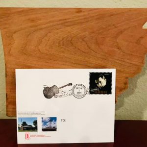 Square postcard stamp of white man text "Johnny Cash" in top right corner silhouette of guitar in middle and two images of post office building in lower left side propped up against wooden board in shape of Arkansas state