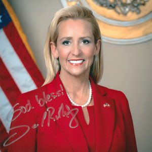 Autographed picture middle aged white woman with long hair wearing suit smiling standing in front of American flag and Arkansas seal inscription "God bless!"