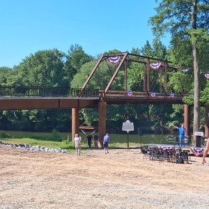 Iron bridge covered in round flags with stars and stripes next to white man on stage speaking into microphone in front of empty folding chairs people walking nearby