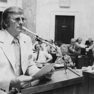 White man wearing suit floral tie and tinted glasses standing at lectern with microphone holding papers in both hands group of people seated in rows in background