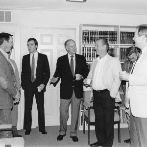 Seven white men dressed in suits holding paper cups standing around older white man talking in office building