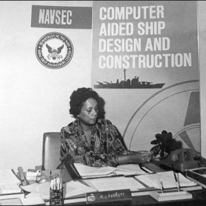 Black woman with short hair sitting at desk filled with documents looking down under sign text "NAVSEC Computer Aided Ship Design and Construction"