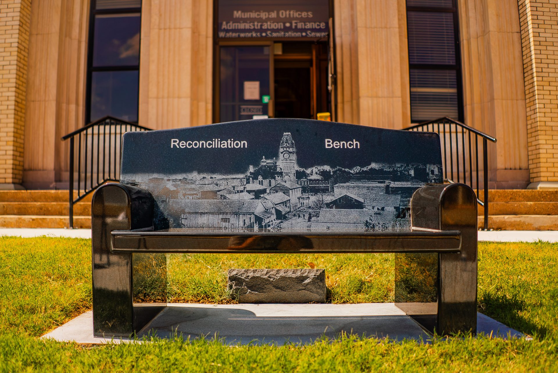 Marble bench with engraved illustration of street scene text "Reconciliation Bench" with entrance into Municipal office building in background