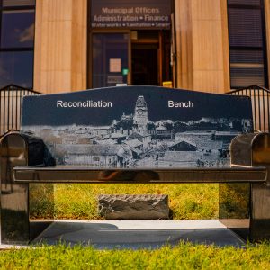 Marble bench with engraved illustration of street scene text "Reconciliation Bench" with entrance into Municipal office building in background