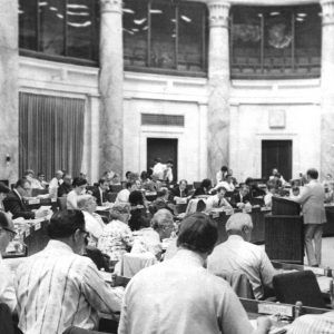 Large group of people seated in semi circle table arrangements all facing white man dressed in tan suit standing at lectern