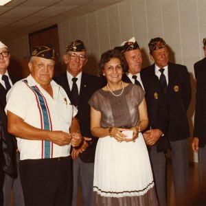 White woman wearing dress and pearl necklace standing with seven white men six wearing suits and ties all wearing American Legion membership caps