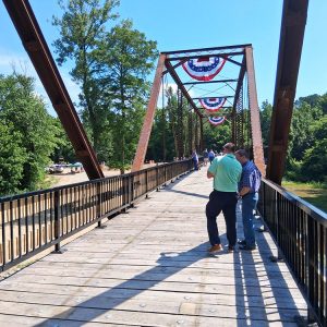 People standing and walking across iron bridge with wooden walkway platform round flags with stars and stripes lined on top